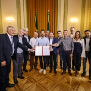 Uma foto de um grupo de treze pessoas (doze homens e uma mulher) em um salão formal, celebrando a autorização de obras. O grupo está posicionado em arco, sorrindo para a câmera, com o governador Eduardo Leite no centro.

Eduardo Leite (de camisa social azul-clara, calças claras e tênis brancos) está segurando um documento aberto (possivelmente a autorização ou convênio) com outros seis homens que também estendem as mãos para tocar a pasta aberta. Os homens vestem camisas sociais, ternos ou blazers. A única mulher no grupo está à direita, vestindo uma calça escura e uma blusa branca com ombros estruturados.

O ambiente é luxuoso, com um piso de madeira em parquet com padrão geométrico de losangos. O fundo tem paredes verde-claras com detalhes em moldura dourada e, no centro, duas bandeiras (a do Brasil e a do Rio Grande do Sul) em mastros altos, emolduradas por cortinas douradas. Em primeiro plano, no canto inferior esquerdo, parte de um celular é visível sendo segurado pela mão de alguém que tira uma foto.
