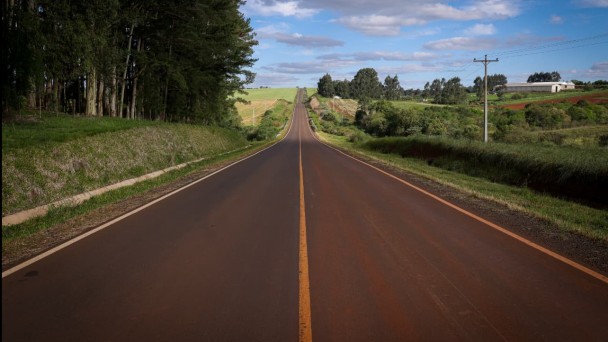 A imagem mostra uma estrada asfaltada, reta e longa, com uma faixa amarela contínua no centro. A estrada segue em linha reta até o horizonte, passando por pequenas elevações e descidas. sobre um fundo azul claro, iluminado pela luz do dia.