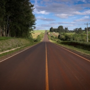 A imagem mostra uma estrada asfaltada, reta e longa, com uma faixa amarela contínua no centro. A estrada segue em linha reta até o horizonte, passando por pequenas elevações e descidas. sobre um fundo azul claro, iluminado pela luz do dia.