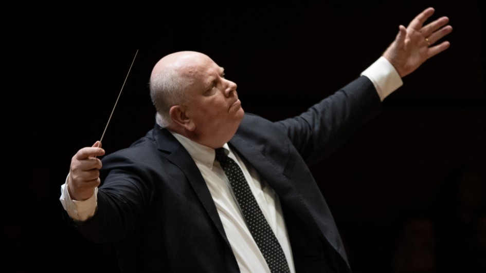 Foto em plano americano de homem de terno com os braços abertos regendo uma orquestra, em fundo escuro. Com a mão direita ele segura a batuta. 