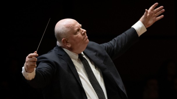 Foto em plano americano de homem de terno com os braços abertos regendo uma orquestra, em fundo escuro. Com a mão direita ele segura a batuta. 