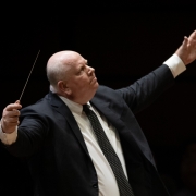 Foto em plano americano de homem de terno com os braços abertos regendo uma orquestra, em fundo escuro. Com a mão direita ele segura a batuta. 