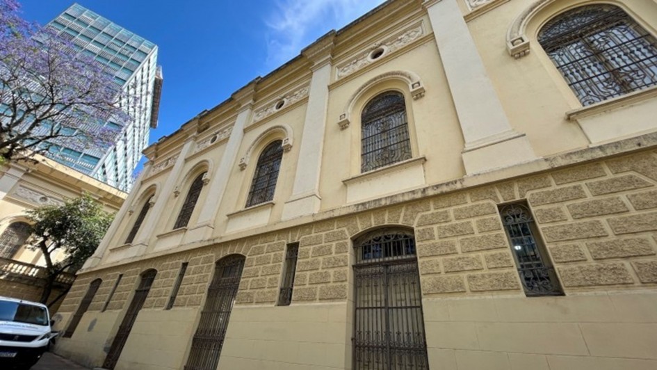 A imagem mostra a fachada de um prédio histórico, com arquitetura clássica e paredes em tom bege. As janelas são altas e arqueadas, protegidas por grades ornamentadas de ferro. A parte inferior da construção apresenta revestimento em pedra trabalhada, enquanto a parte superior exibe ornamentos e detalhes decorativos. À esquerda, parte de uma árvore florida aparece ao lado de um edifício moderno de vidro, criando contraste entre o antigo e o contemporâneo. O céu está azul com poucas nuvens.