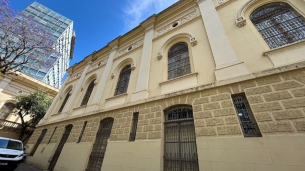 A imagem mostra a fachada de um prédio histórico, com arquitetura clássica e paredes em tom bege. As janelas são altas e arqueadas, protegidas por grades ornamentadas de ferro. A parte inferior da construção apresenta revestimento em pedra trabalhada, enquanto a parte superior exibe ornamentos e detalhes decorativos. À esquerda, parte de uma árvore florida aparece ao lado de um edifício moderno de vidro, criando contraste entre o antigo e o contemporâneo. O céu está azul com poucas nuvens.