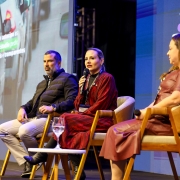 A foto mostra três pessoas sentadas em um palco. A do meio está falando ao microfone. Ao fundo, há um telão colorido. 