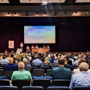 A foto registra uma audiência pública em um auditório grande e lotado, com fileiras de cadeiras ocupadas. No palco, há uma mesa com integrantes da apresentação e um telão exibindo o título “Programa de Parcerias e Concessões – Bloco 1 de Rodovias”. À esquerda, uma pessoa fala ao microfone em um púlpito. O público preenche todo o espaço visível, demonstrando ampla participação no evento.