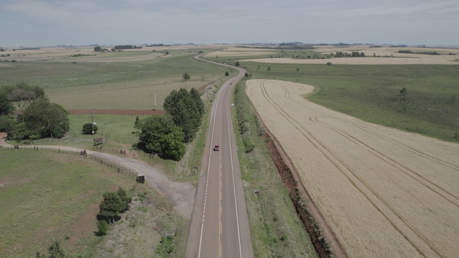 Foto de uma estrada de mão dupla reta, com curvas mais à frente, no meio de área rural.