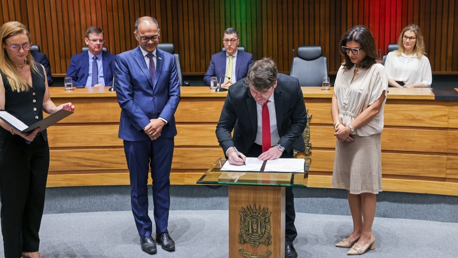 A cena mostra uma cerim&ocirc;nia formal em um audit&oacute;rio. No centro, um homem de terno preto e gravata vermelha assina um documento sobre uma mesa de vidro com um bras&atilde;o na frente. Ao redor dele, tr&ecirc;s pessoas em p&eacute; observam: um homem de terno azul e duas mulheres, uma segurando uma pasta aberta. Ao fundo, outras pessoas sentadas em uma mesa grande de madeira acompanham o ato. O ambiente &eacute; solene, com ilumina&ccedil;&atilde;o suave e painel de madeira atr&aacute;s.