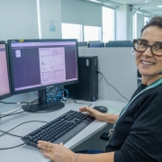 A imagem mostra uma consultora do Telessaúde sentada em frente a sua mesa de trabalho. Ela olha para a câmera e, na sua frente, há dois monitores de computador. 