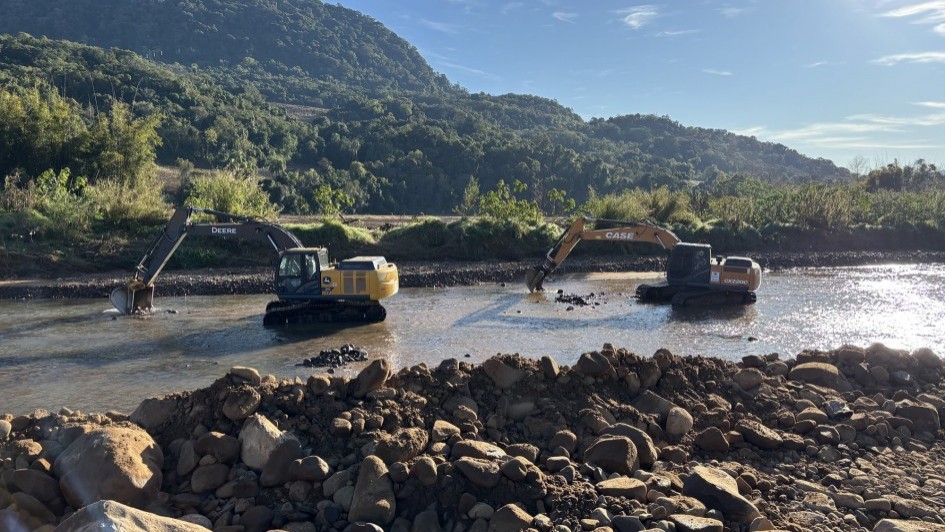 A imagem mostra dois tratores de escavação operando dentro de um rio. Na frente da imagem há um monte de pedras e terra. O cenário inclui uma paisagem montanhosa com vegetação densa no fundo.