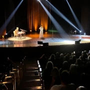 Apresentação ao vivo em teatro com duas pessoas no palco, luzes dramáticas e público atento nas cadeiras. Atmosfera artística e envolvente.