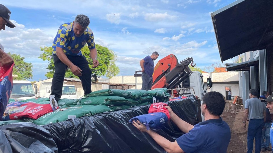 Pessoas descarregam sacos de produtos de caminhão com lona preta em área rural. Trabalho em equipe e esforço físico.