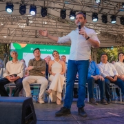 Homem fala ao microfone em palco coberto, com público sentado ao fundo. Evento formal e comunitário.