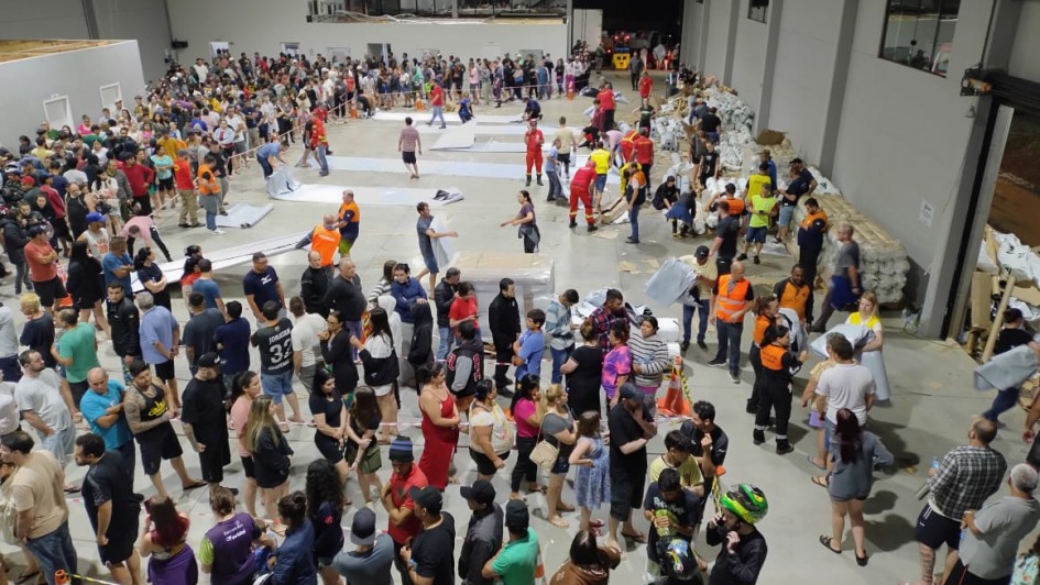 Imagem mostra um grande salão com muitas filas de pessoas aguardando serem atendidas. Auxiliares da Defesa Civil com coletas laranjas auxiliam em diversos pontos do salão.