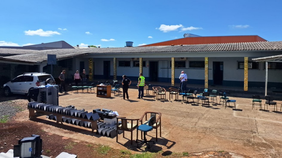 A imagem mostra o pátio externo de uma escola, com diversas cadeiras e equipamentos organizados ao ar livre. Algumas pessoas caminham e inspecionam o local. O prédio escolar ao fundo apresenta estrutura simples, com telhado de duas águas e portas distribuídas ao longo da fachada. O céu está limpo e ensolarado.