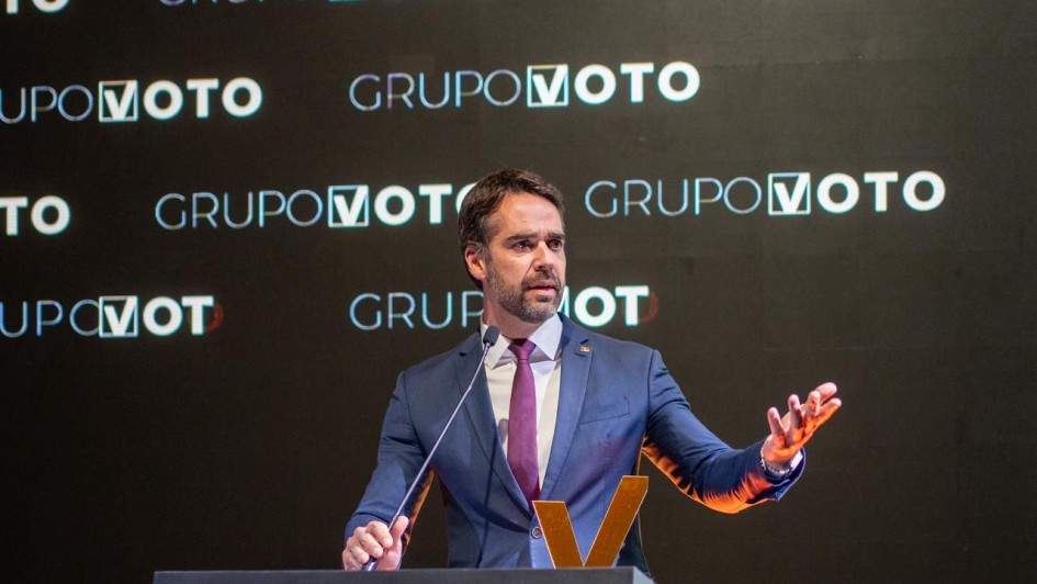 Um homem, presumivelmente o Governador Eduardo Leite, está em pé atrás de um púlpito de cor escura, fazendo um discurso. Ele veste um terno azul-marinho, uma camisa branca e uma gravata roxa escura. Ele tem barba e cabelo curto e escuro. A mão direita dele está apoiada sobre o púlpito, e a mão esquerda está levantada ao lado, com a palma aberta, como se estivesse gesticulando enquanto fala. O fundo é escuro, e as palavras "GRUPO VOTO" estão visíveis repetidamente em letras maiúsculas brancas atrás e acima dele. Um grande 'V' estilizado em dourado ou laranja é visível na parte frontal do púlpito. A iluminação está focada no orador, tornando o fundo menos distinto.