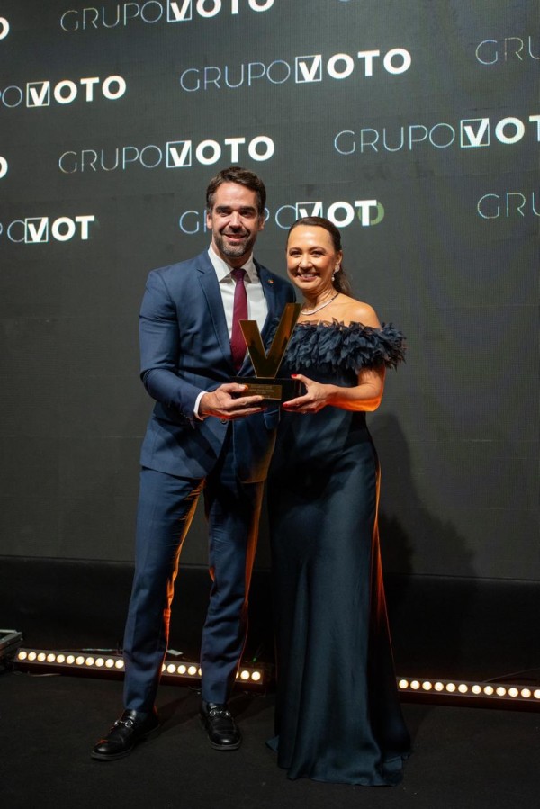 A fotografia mostra um homem (presumivelmente o Governador Eduardo Leite) e uma mulher em pé lado a lado, sorrindo e segurando um prêmio.

O homem está à esquerda, vestindo um terno azul-marinho, camisa branca e gravata bordô/vermelha. Ele tem cabelo curto, escuro e barba.

A mulher está à direita, vestindo um longo vestido de cor azul-marinho ou chumbo, com detalhes de penas ou babados no ombro e decote. Ela tem cabelo escuro preso e usa brincos de argola.

Ambos estão segurando juntos um troféu grande, estilizado com a forma da letra 'V' (remetendo ao GRUPO VOTO), na cor dourada ou bronze.

O fundo é uma parede ou tela escura, na qual o logotipo e o nome 