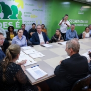 Foto de uma sala de reunião,  em que aparecem mais de 20 pessoas sentadas. Elas vestem trajes formais. Enquanto algumas conversam, outras observam. 