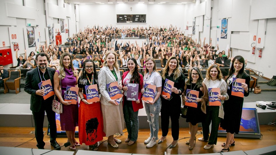 A foto mostra um audit&oacute;rio lotado, com estudantes e participantes levantando as m&atilde;os ao fundo. Na parte frontal do palco, um grupo de mulheres e homens posa para a foto segurando &aacute;lbuns de figurinhas da Lei Maria da Penha. Todos est&atilde;o alinhados em uma fileira, sorrindo para a c&acirc;mera. O espa&ccedil;o &eacute; amplo, bem iluminado, com cadeiras ocupadas ao fundo e estrutura de audit&oacute;rio tradicional.