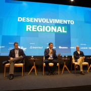 Foto de palco de cor escura com telão azul ao fundo. Quatro homens aparecem sentados. Um deles fala ao microfone.