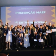 A imagem registra um grande grupo de pessoas no palco, celebrando um evento de premiação.

Pessoas e Ação: Cerca de 15 a 20 pessoas (homens e mulheres) estão no palco, posando juntas. A maioria está vestindo roupas casuais a semi-formais e acessórios (como crachás de evento). Muitas pessoas estão levantando os braços e as mãos em sinal de celebração e entusiasmo. Algumas seguram certificados ou documentos.

Fundo (Tela de Projeção): A tela de projeção, que domina o fundo, exibe o tema do evento.

No topo, lê-se: "5º Mercado Audiovisual Entre Fronteras" e, em uma linha menor, o slogan: "Onde as fronteiras se diluem, as histórias se encontram".

No centro, em letras grandes e em negrito: "PREMIAÇÃO MAEF" (sendo MAEF a sigla do Mercado Audiovisual Entre Fronteras).

Abaixo, há o logotipo estilizado do "5º MERCADO AUDIOVISUAL ENTRE FRONTERAS".

Ambiente: A cena se passa em um ambiente interno, provavelmente um auditório ou salão de eventos. O palco é elevado e há caixas de som visíveis nas laterais. A iluminação é focada no palco.

Outros Detalhes: No lado direito, parte de um banner promocional do evento "ENTRE FRONTERAS" é visível, repetindo a identificação do evento. No canto inferior direito, o crédito do fotógrafo "crédito Sérgio Rosa" é mencionado no nome do arquivo.

Tema Geral: A imagem é um retrato de grupo no palco durante a cerimônia de premiação do 5º Mercado Audiovisual Entre Fronteras, mostrando a alegria e celebração dos participantes ou vencedores.