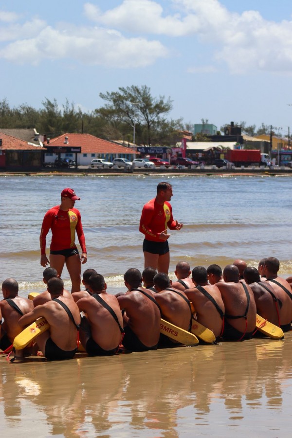 A foto mostra um grupo de guarda-vidas sentados na areia da praia, organizados em fileira, voltados para dois instrutores que estão em pé à frente, próximos à água.