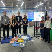 Foto posada de oito pessoas em ambiente fechado, numa sala de exposição no simpósio. A pessoas estão de pé e usam crachá, e encontram-se próximas a uma mesa baixa com livros e folhetos e a uma TV ao fundo.