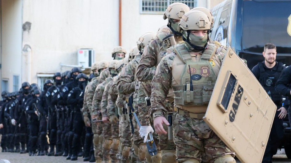 Uma foto em plano médio de seis pessoas (quatro mulheres e dois homens) posando juntas no momento da assinatura de um convênio ou portaria em Cruzeiro do Sul. Elas estão em frente à entrada de um prédio moderno e recém-reformado (possivelmente uma Unidade de Referência em Saúde - URS).

A mulher no centro, de blusa e blazer azul-claro sobre calça escura, segura o documento assinado. Ela é ladeada por cinco outras pessoas que estendem as mãos em direção ao documento, sorrindo. O homem ao lado esquerdo dela veste uma camisa polo bege, e o homem ao lado direito veste uma camisa social azul-clara. A mulher na extrema direita está vestida com um longo vestido rosa vibrante e um colar de miçangas coloridas.

O cenário é em frente a um edifício de dois andares com fachada branca e azul-clara. Uma escadaria de acesso com corrimões brancos está visível à direita. No canto esquerdo, há mastros com as bandeiras do Brasil, do Rio Grande do Sul e de Cruzeiro do Sul. O grupo está de pé sobre um tapete ou plataforma escura. O evento ocorreu ao ar livre, no final da tarde ou começo da noite, com boa iluminação frontal.