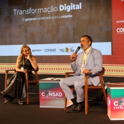 Três painelistas estão sentados em cadeiras, com cubos coloridos entre eles, com o nome do evento. O mediador, ao centro, está falando ao microfone. À esquerda dele na foto, há uma painelista mulher, loira, com vestido marrom, que o olha atentamente. Do outro lado, um homem, de terno claro, sorri olhando para o papel. 