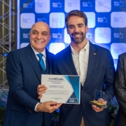 Três homens posam para a foto segurando um certificado e uma pequena estatueta.
No centro está o governador Eduardo Leite, sorrindo e segurando o certificado ao lado de dois representantes da Santa Casa de Pelotas. O fundo exibe um painel institucional azul e branco.