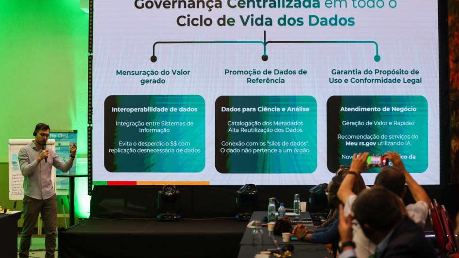 Foto mostra painelista em pé fazendo a apresentação, em frente a uma tela com gráficos sobre ciclo de vida dos dados. Algumas pessoas estão sentadas e tirando foto da tela. 