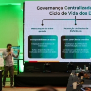 Foto mostra painelista em pé fazendo a apresentação, em frente a uma tela com gráficos sobre ciclo de vida dos dados. Algumas pessoas estão sentadas e tirando foto da tela. 