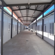 Uma foto de um corredor coberto (passarela ou área de circulação) em um complexo de edifícios modulares (contêineres ou salas pré-fabricadas), possivelmente uma escola. O corredor é longo e estreito, com simetria acentuada.
As paredes laterais dos prédios são brancas. As janelas, também brancas, possuem grades de proteção verticais e horizontais. Portas brancas de entrada para as salas estão visíveis ao longo de ambos os lados do corredor.
A estrutura da cobertura é sustentada por vigas escuras (pretas ou marrom-escuras) de metal, formando um telhado inclinado. O piso do corredor é de cimento. A iluminação é fornecida por luminárias brancas de LED fixadas na parte inferior do telhado. A luz natural entra pela abertura superior da cobertura, e no fundo, é visível uma área de pátio e construções adjacentes.