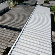 Uma foto aérea com ângulo inclinado, mostrando o telhado de um complexo de edifícios, possivelmente uma escola. A imagem destaca um grande telhado novo, de metal cor cinza-claro e formato ondulado, que cobre uma longa estrutura retangular no centro da foto.
Este telhado novo contrasta com os telhados antigos ao redor. Os telhados adjacentes são de amianto (ou material similar), de cor cinza escura e com marcas de tempo e sujeira. A luz do sol incide sobre o telhado novo, refletindo a cor clara.
Ao redor das estruturas, há pequenas áreas de pátio cimentado e árvores. A imagem sugere a revitalização ou reforma da cobertura de uma parte do complexo escolar.