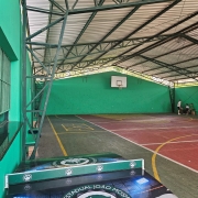 Uma foto de uma quadra poliesportiva coberta (possivelmente de um colégio estadual) com foco em uma mesa de tênis de mesa (pingue-pongue) em primeiro plano.
Em primeiro plano:
A mesa de tênis de mesa tem uma superfície de jogo verde-escura e um design no corpo principal (a lateral da mesa).
O design da mesa apresenta o nome "COLÉGIO ESTADUAL JOÃO MOSMANN" em arco, circundando um emblema ou logotipo com as iniciais "C E J M" e o mapa do Rio Grande do Sul.
A iluminação azul e verde é projetada artificialmente no design da mesa.
A quadra:
A quadra é coberta por um telhado de metal sustentado por uma estrutura metálica de treliças escuras (verde-escuras ou pretas).
O piso da quadra é vermelho e verde (com divisões amarelas) e exibe marcações para esportes (o círculo central é visível).
A parede de fundo é de cor verde-água/turquesa vibrante e apresenta uma tabela de basquete branca com aros.
No canto direito, dois jovens estão sentados em um banco ou área de descanso.
A estrutura lateral da quadra (à esquerda) é gradeada com barras verticais. A foto sugere uma área de lazer e esportes de uma instituição de ensino.