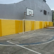 Uma foto da fachada de um prédio escolar sob um céu azul-claro com nuvens. A fachada do edifício de dois andares (vista lateral em ângulo aberto) é de cor cinza-claro.
O que se destaca é a aplicação de painéis retangulares coloridos entre as janelas de cada andar, em uma sequência vibrante:
Um painel em vermelho.
Um painel em verde.
Um painel em amarelo.
Um painel em verde-musgo.
Um painel em laranja/vermelho queimado.
Um painel em verde-oliva.
As janelas são múltiplas janelas-guilhotina (tipo basculante) com grades de metal finas (quadriculadas) e escuras. O telhado é cinza escuro. À esquerda da construção, há árvores frondosas, e na base do prédio, em primeiro plano, há um canteiro de terra e um pequeno caminho cimentado. A revitalização utiliza cores para dar um aspecto moderno e alegre à escola.