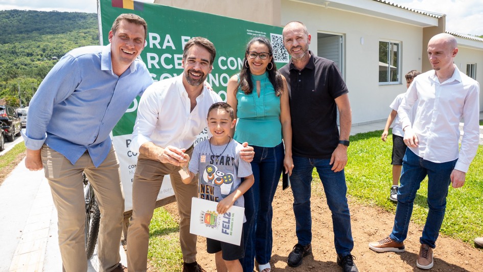 O governador e uma família posam para a foto em frente a uma casa entregue. Eles sorriem e uma criança segura a chave da moradia.