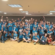 Uma foto externa e panorâmica de uma grande delegação gaúcha (cerca de 50 a 60 pessoas, de todas as idades) reunida em uma sala de conferências ou salão de hotel. O grupo parece ser a delegação para as Paralimpíadas de 2025.

As Pessoas:

A maioria está vestindo camisetas polo azuis-turquesa com detalhes amarelos nos ombros e no colarinho, que são os uniformes da delegação.

Os membros do grupo estão sentados no chão, agachados ou em pé, e muitos levantam os braços e acenam, sorrindo de forma entusiasmada.

Diversos atletas em cadeiras de rodas estão visíveis na primeira fileira e no centro da imagem.

O Ambiente:

A sala tem iluminação clara vinda de luminárias embutidas no teto em padrões retangulares.

O chão é acarpetado com um padrão geométrico em tons de amarelo e marrom-claro.

No canto esquerdo, algumas cadeiras azuis de plástico estão empilhadas.

A cena transmite um clima de união, celebração e entusiasmo.