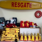 A imagem mostra uma sala com diversos equipamentos de salvamento utilizados por guarda-vidas. Na parede, há uma prancha de resgate amarela com a palavra “RESGATE” em vermelho. Ao lado, está pendido um flutuador circular vermelho. Sobre uma mesa, encontram-se mochilas vermelhas, bolsas de primeiros socorros, cilindros de oxigênio, colar cervical, protetor solar e outros itens médicos. No chão, empilhados, há vários flutuadores amarelos com a inscrição “CBMRS – GUARDA-VIDAS”, além de pares de nadadeiras também nas cores amarela e vermelha embaladas em plástico. À esquerda, aparecem pás e outros materiais de apoio.