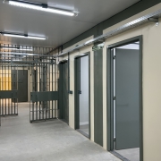 A imagem é uma fotografia de um corredor interno da Casa de Custódia da Polícia Civil em Porto Alegre, focada em um ambiente que combina elementos de segurança institucional com uma infraestrutura nova ou reformada.

Elementos da Cena:
Estrutura de Segurança (Esquerda e Fundo):

No lado esquerdo e no fundo, há grades metálicas escuras (celas ou portões de segurança), típicas de áreas de custódia.

Um portão de grade está aberto, revelando um interior com paredes amarelas/alaranjadas escuras.

Corredor e Paredes (Direita):

O corredor principal possui um piso de cimento cinza claro e paredes de cor bege claro/amarelado.

Há portas fechadas de cor cinza-escura ao longo da parede direita, indicando possíveis salas ou celas individuais. Uma das portas está ligeiramente aberta, revelando um interior de cor clara, possivelmente um banheiro.

As portas têm batentes e detalhes verticais de cor cinza-escura, contrastando com a parede.

Infraestrutura e Iluminação:

O teto é baixo e claro, equipado com luminárias de LED tubulares (fluorescentes) que fornecem iluminação branca e intensa.

Na parte superior das paredes e teto, há calhas metálicas (eletrocalhas) expostas, indicando a passagem de cabos elétricos ou de dados, o que sugere uma instalação moderna ou recente.

Há também uma placa de sinalização de saída de emergência ("SAÍDA") verde e branca acima de uma das portas.

Conclusão Visual:
A foto representa a área interna de custódia, destacando a combinação de elementos de segurança (grades) com uma infraestrutura moderna (iluminação e cabeamento exposto), possivelmente resultado de uma obra de construção ou reforma.