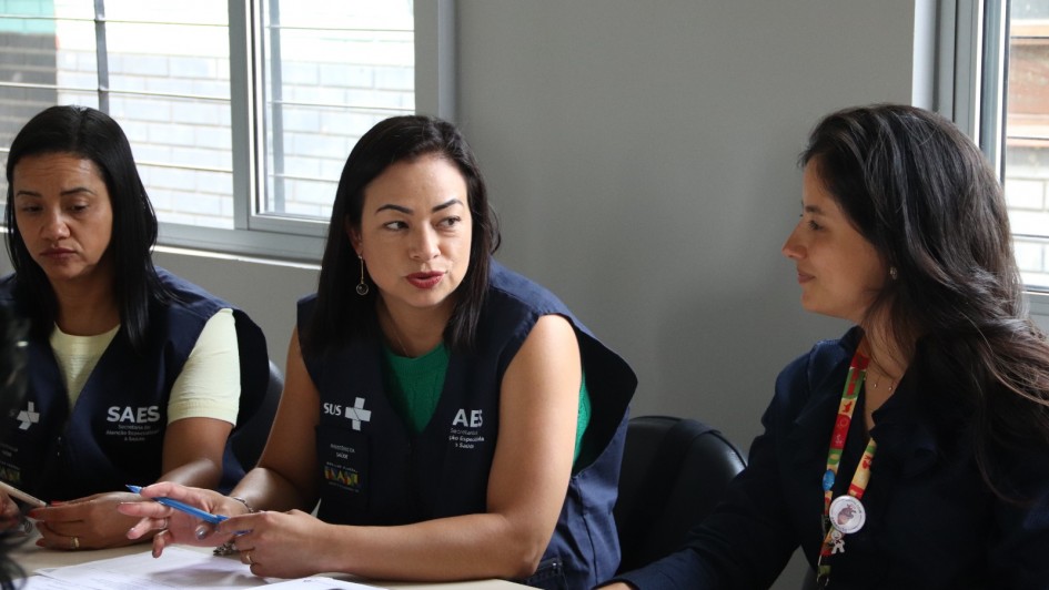A imagem mostra três mulheres sentadas lado a lado, em uma mesa de trabalho, vestindo coletes com inscrições do SUS e SAES. Elas conversam entre si e seguram canetas e papéis. 