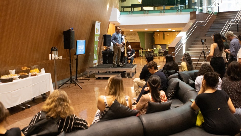 A imagem mostra um evento realizado em um espaço interno amplo, com piso de granito e escadaria lateral. Em um pequeno palco, um homem fala ao microfone para o público sentado em sofás e cadeiras. À esquerda, há uma mesa com coffee break. Ao fundo, aparece um banner da Secretaria da Cultura do Rio Grande do Sul.