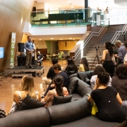 A imagem mostra um evento realizado em um espaço interno amplo, com piso de granito e escadaria lateral. Em um pequeno palco, um homem fala ao microfone para o público sentado em sofás e cadeiras. À esquerda, há uma mesa com coffee break. Ao fundo, aparece um banner da Secretaria da Cultura do Rio Grande do Sul.