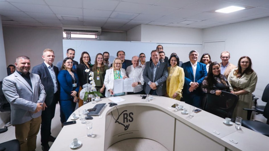 Um grupo numeroso de pessoas está em pé, reunido para a foto, atrás de uma mesa em formato de U. Ao centro, a secretária da Saúde segura o documento assinado. 