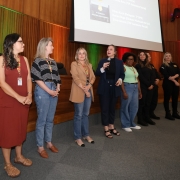 Foto meio de lado de 7 mulheres e 1 homem à frente de um auditório. Eles estão de pé e, ao centro, uma mulher fala ao microfone.