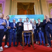 A foto mostra um grupo de aproximadamente 15 pessoas reunidas em um palco com carpete vermelho, dentro de um salão amplo com colunas claras e iluminação em tons de roxo e amarelo. Ao centro, o governador Eduardo Leite, segurando um documento aberto com ambas as mãos, posicionado sobre uma mesa pequena e estreita. As pessoas ao redor batem palmas. Atrás do grupo, há um grande painel digital exibindo cores em azul e verde.