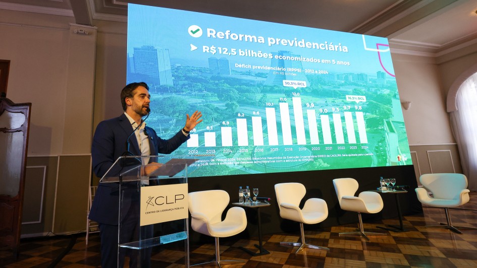 Foto de Leite falando ao púlpito com o braço esquerdo erguido à altura do ombro, apontando para o telão atrás, no qual se lê Reforma Previdenciária, com um gráfico logo abaixo.