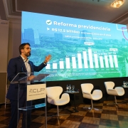 Foto de Leite falando ao púlpito com o braço esquerdo erguido à altura do ombro, apontando para o telão atrás, no qual se lê Reforma Previdenciária, com um gráfico logo abaixo.