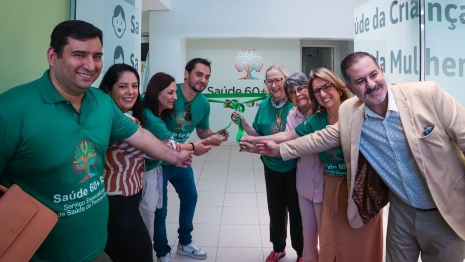 Um grupo de oito pessoas está alinhado em um corredor interno, posicionado em semicírculo, todas estendendo as mãos para o centro para segurar uma fita verde, como parte de uma cerimônia de inauguração. Várias pessoas usam camisetas verdes com o logotipo do programa “Saúde 60+ RS”, que apresenta uma árvore estilizada multicolorida. À esquerda da imagem, um homem segura uma pasta bege. As portas de vidro abertas ao fundo têm adesivos com textos relacionados a serviços de saúde, como “Saúde da Criança” e “Saúde da Mulher”.
