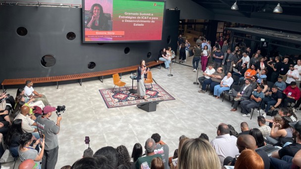 Foto mostra visão de cima de um auditório onde uma mulher apresenta diante de uma plateia, com um telão mostrando uma programação na tela. 